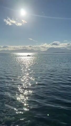 The video shows a wide shot of a body of water with a bright sun in the upper left corner. The sun's reflection creates a glittering path across the water's surface. In the distance, there appears to be a small boat with people in it. The horizon is lined with mountains or land formations under a partly cloudy sky. The water itself is a deep blue, with gentle ripples. The overall scene is serene and appears to be set during daytime.