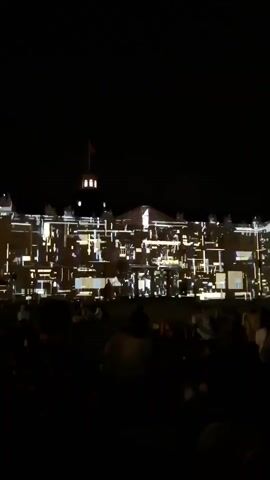 The video shows a nighttime outdoor event with a crowd of people sitting on the grass facing a large building. The building is illuminated with colorful lights and projections. The projections on the building change throughout the video, from abstract colorful patterns to geometric shapes resembling windows. The crowd appears to be watching the light show.
