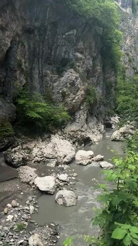 A video displays a rocky gorge with a river flowing through it. The river is murky and appears to be flowing swiftly, with several large rocks visible in and around it. The gorge walls are steep and covered in patches of green vegetation. The light suggests it is daytime, possibly overcast.