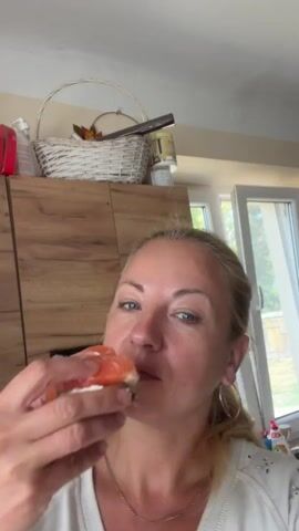 A woman is shown from the chest up, with her head turned slightly to her left, eating a piece of food. She has blonde hair tied back, and is wearing a white top and hoop earrings. She is holding a piece of food between her fingers, and her mouth is open as she bites into it. The food appears to be a piece of salmon on a white base, possibly sushi or a sandwich. Her eyes are closed as she chews. The background is blurred but appears to be a kitchen, with wooden cabinets and a basket on top of them. A window is visible to the right, with white curtains. The lighting suggests it is daytime.