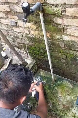 A person is shown cleaning fish under a running faucet attached to a brick wall. The person is holding a fish in their left hand and a knife in their right hand, scraping the scales off the fish. Another fish is visible on the ground next to the person's right leg. The environment appears to be outdoors, with a weathered brick wall and a concrete surface that is damp and has some moss growth. A PVC pipe runs vertically along the wall.