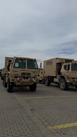 The video displays a static shot of several military Light Medium Tactical Vehicles (LMTV) in a desert tan color, parked outdoors on a paved surface. The camera slowly pans from left to right, revealing more trucks.

In the foreground, a prominent LMTV is parked on the left, facing the camera. Text visible on its front bumper includes "90100" and "0121." A small sticker or text "V7JK83" is visible above its windshield, along with a white sticker on the windshield itself.

To the right of the foreground truck, two more LMTV trucks are parked in a row, facing the same direction. The one immediately to the right has a large, enclosed cargo box on its bed. Text "41-2-3028" is visible on its front bumper. Other similar trucks are visible further in the background.

The trucks are parked on a gray-toned, patterned paved surface, possibly interlocking bricks, with yellow painted lines on the ground, indicating parking spaces.

The sky is partly cloudy, with a mix of overcast grey and patches of blue sky, suggesting daytime. There are no people visible in the video. The scene appears to be a military staging area or a parking lot on a base in Illesheim, Germany.