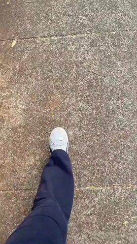 The video begins with a close-up of a person's feet in white sneakers walking on a concrete path. The view then pans up to show a sidewalk next to a road lined with trees. Houses are visible in the distance. The person continues walking on the sidewalk.