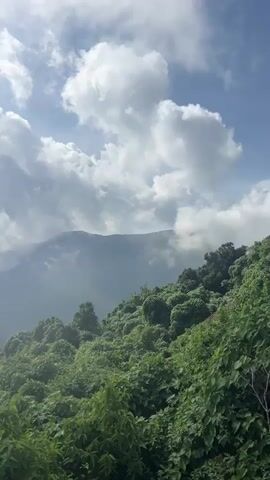 The video captures a time-lapse of a mountainous landscape under a dynamic sky. Lush green trees and foliage cover the foreground and midground slopes. In the background, mountains are partially visible, intermittently obscured by rapidly moving white and grey clouds that roll over the peaks and across the sky. The clouds appear to be moving from left to right, creating a sense of motion and change in the weather. The sky above is a mix of bright blue and white clouds, suggesting a clear yet active day. There are no people or specific objects in the frame, focusing entirely on the natural environment.