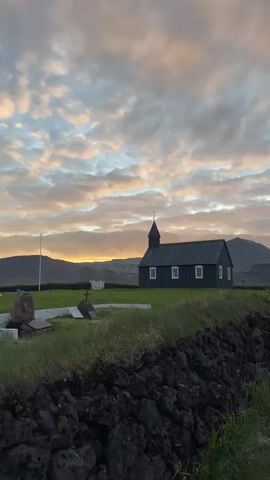 The image presents a serene rural landscape in Búðir, Iceland, centered around the iconic black Búðakirkja church. The small, dark-colored wooden church stands prominently in a green grassy field, featuring a distinct steeple, a dark roof, and several white-framed windows on its side. To the left of the church, a white flagpole is partially visible, and in the foreground, several gravestones or cemetery markers are scattered within the grass, indicating a churchyard. A low, rugged wall made of dark, possibly volcanic rocks, occupies the immediate foreground, adding texture to the scene.

The background is dominated by rolling hills or mountains, which recede into the distance under a vast, expansive sky. The sky itself is a striking feature, filled with a mix of soft, diffused clouds displaying hues of warm orange, pink, and light grey, suggesting either a tranquil sunrise or sunset. This soft, natural light bathes the entire scene, creating a peaceful and somewhat ethereal atmosphere.

No people are visible in the photograph, and no specific activity is taking place, emphasizing the solitude and natural beauty of the remote setting. The weather appears calm, with the clouds providing a dramatic backdrop without obstructing the view. There is no visible text in the image. The overall impression is one of quiet contemplation and the enduring presence of history within a stunning natural environment, characteristic of the unique Icelandic landscape.