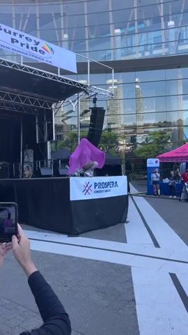 A person dressed in a pink shroud with white sneakers and silver detailing is dancing on a stage. The stage has a black curtain backdrop and a banner that says "PROSPERA CREDIT UNION." In the background, a modern glass building reflects the sky and other buildings. To the left, a banner with "Surrey Pride" and a rainbow flag is visible. To the right, a pink tent is set up with people standing near it. A person in the foreground is holding a phone, recording the performance. The event appears to be outdoors, with a paved surface in front of the stage.