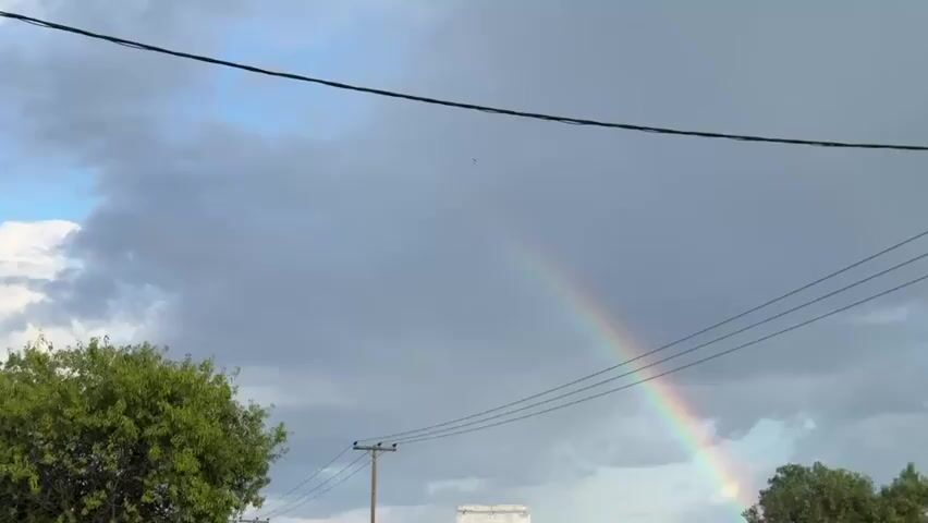 The video shows a rainbow in the sky. It is a full rainbow with all colors visible, but it is not very bright. The sky is cloudy and there are power lines running across the view. There are also trees visible in the foreground. A car passes by on the road.