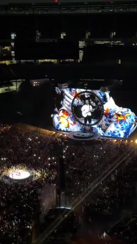 The video is an aerial shot of a large outdoor concert at night. A massive screen displays a circular image of a male performer holding a guitar, with abstract colorful graphics surrounding him. Below the screen, a vast crowd of concertgoers holds up their phones, their lights creating a sea of small lights. A circular stage is visible in the lower left quadrant, with a few figures on it. The overall atmosphere is dark, with stage lighting illuminating the performer and the screen. The background shows the dark structures of a stadium or venue.
