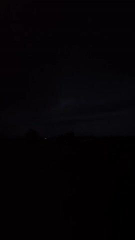 This media file captures a very dark, possibly stormy, night scene over a flat landscape. The image is predominantly black with very subtle details visible. Silhouetted against the slightly less dark sky are faint outlines of what appear to be trees and possibly some distant structures or lights. The ground in the foreground is entirely black, obscuring any features. The sky is dark and appears overcast, hinting at night or a heavily clouded day. There are no discernible people or specific objects. The setting suggests an outdoor, rural or undeveloped area. The overall mood conveyed is one of darkness and potential obscurity, with no clear activity or event taking place. The time of day is clearly night. There is no visible text.