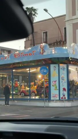 A car drives through a European town. The car passes a small, colorful fairground ride for children, decorated with cartoon characters.  The car passes a motorcycle parked on the sidewalk, followed by an older, pink building with a small church in the distance. There are several cars and pedestrians on the street.