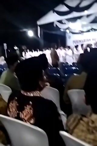 The video captures an outdoor nighttime event with a stage set up under a tent. A banner on the stage reads "PERINGATAN HUT RI KE-81," indicating an 81st anniversary celebration of the Republic of Indonesia. A group of people, mostly wearing white, are standing on the stage, possibly performing or being recognized. In the foreground, an audience sits in rows of white chairs, facing the stage. Some audience members are wearing traditional Indonesian attire, including batik shirts and peci hats. The lighting appears to be from stage lights and perhaps some ambient streetlights. The atmosphere seems formal and celebratory.