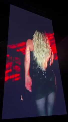 The video begins with a shot of a large screen displaying a blonde woman with long, wavy hair in a black outfit. The camera then pans out to show the stage setup, which includes a large circular backdrop with a crown logo inside. Red lights illuminate the stage structure and the logo. A band performs on stage in front of a large crowd. The environment is a concert venue at night.