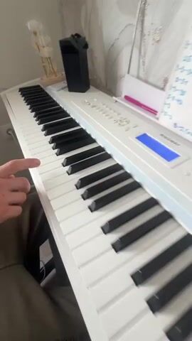 The video shows a close-up of a person's hands playing a white digital piano. The camera is positioned above and to the side of the piano keyboard, focusing on the right hand of the person as they press down on the keys. The fingers are visible, pressing several white keys in sequence. The piano has black and white keys, and some of the controls and a music stand are visible in the background. The lighting suggests an indoor setting, possibly a home or studio. There are decorative objects, including what appear to be dried flowers in a vase, on top of the piano. The person is wearing a khaki-colored garment on their lower body. The overall scene depicts a moment of musical practice or performance.