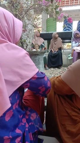A group of people, mostly women wearing hijabs, are gathered outdoors. Some are sitting on the ground, while others are standing. There are trees with pink flowers and buildings in the background. A motorcycle is parked nearby. The camera is held low and is angled upwards, focusing on the people and the surrounding environment. The scene appears to be a community gathering or event.