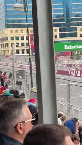 The video shows a Formula 1 race car passing by a crowd of people in a stadium. The car is pink and blue, and is speeding along a gray track. The crowd is standing behind a gray metal barrier, and they are all looking at the car. Behind the barrier and the race track are buildings, and advertisements on the edge of the race track. The sky is overcast.