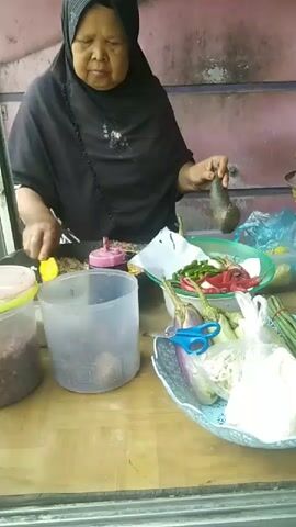 An older woman wearing a black hijab and black clothing is standing behind a wooden counter, preparing food. She is holding a yellow utensil in her right hand and appears to be stirring something in a dark pan. To her left is a clear plastic container with a pink lid, and next to it is a larger, clear plastic container filled with a dark liquid. In front of her on the counter, there is a white piece of paper with some food items on it, and to the right, a clear plastic bowl filled with various vegetables, including sliced eggplant, green chili peppers, red chili peppers, and other ingredients. There is also a bunch of green beans secured with a rubber band, and a plastic bag containing white food items. A pair of blue scissors rests on the vegetables in the bowl. The background appears to be an outdoor stall or food cart with a pink and gray wall. The lighting is natural.