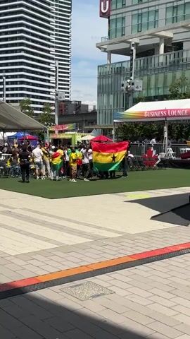 A group of people are gathered on a patch of artificial turf, holding a large Ghanaian flag. The flag is spread out, with some individuals holding the edges and others standing within its confines. The people are dressed in casual clothing, with many wearing yellow shirts, some of which appear to be soccer jerseys. In the background, there are tents, food stalls, and modern buildings. The scene suggests an outdoor event or festival.