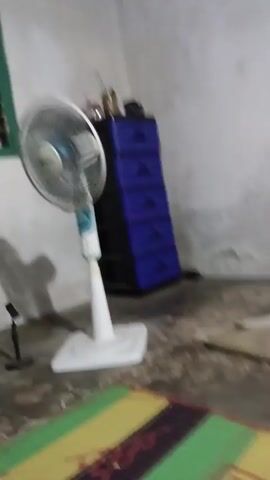 The video shows a still shot of a room. In the center is a white oscillating fan on a square base. To the left of the fan is a black shelf unit with a television on top. To the right of the fan is a blue plastic cabinet with five drawers. The floor is concrete and a colorful mat is visible in the foreground. The walls are white and a window with green shutters is on the left.
