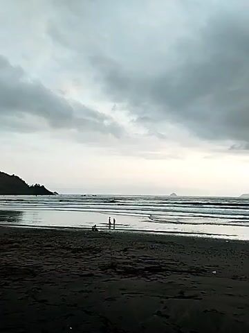 A wide shot captures a gloomy beach with dark sand in the foreground. The ocean stretches out to the horizon, with gentle waves breaking on the shore. In the middle distance, a few people are visible near the water's edge, appearing as small figures against the vastness of the sea and sky. The sky is overcast with heavy, gray clouds, lending a somber mood to the scene. To the left, a lush green hillside rises from the coastline.