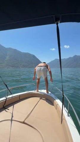 A person stands on the bow of a boat, facing away from the camera, with their arms spread. The boat is on a body of water, with mountains in the background under a clear blue sky. The person then jumps into the water, creating a splash. The camera remains stationary on the boat.