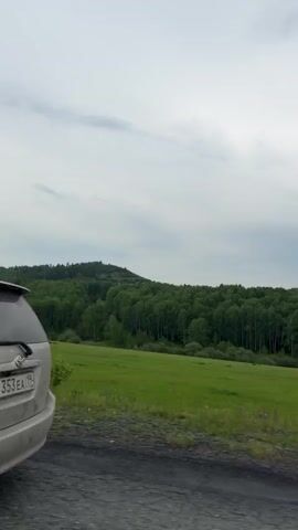 The video shows a side view of a silver car driving on a road. A person is visible in the driver's seat, holding the steering wheel. Outside the car, there is a landscape with rolling green hills covered in trees. The sky is overcast with clouds.
