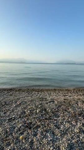 The video shows a close-up view of a rocky shore with the calm, blue water of a lake or sea in the background. The sky is clear and pale blue. The camera is positioned low to the ground, focusing on the pebbles and small debris on the shore. Gentle waves lap at the edge of the water. In the distance, faint landforms or mountains are visible on the horizon. The lighting suggests it is either early morning or late afternoon. There are no people or animals visible in the video.