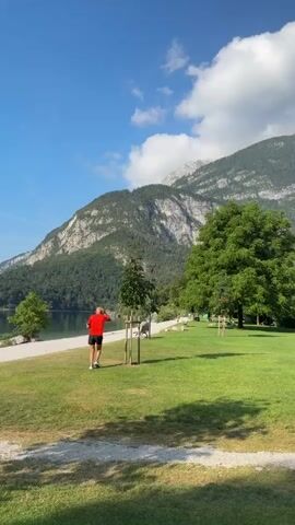 A person in a red shirt and black shorts runs along a paved path next to a body of water. They are surrounded by green grass and trees. In the background, there is a large mountain with some clouds. The sky is blue with white clouds. The scene is well-lit and appears to be a sunny day.