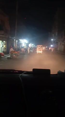 Here is a concise description of the video:

The video shows a night scene from inside a vehicle moving slowly down a street in Pakistan.  The street is lit by streetlights and lights from small businesses lining the road. People are walking and standing around on the sidewalks and near parked motorcycles. The street is relatively empty with only a few other vehicles visible.