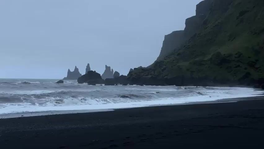 The video depicts a coastal scene featuring a black sand beach under an overcast sky. Waves break along the shoreline, with the surf appearing moderately rough. In the distance, jagged rock formations rise from the water, partially obscured by the atmospheric haze. To the right, a large cliff covered in vegetation dominates the landscape. The overall tone is bleak and dramatic.