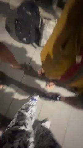 The video shows a group of people's feet on a paved surface. One person is wearing sandals with bright pink nail polish, and another is wearing patterned pants and sneakers. The camera pans up to show the faces of two women, one of whom is laughing. The scene appears to be outdoors at night.