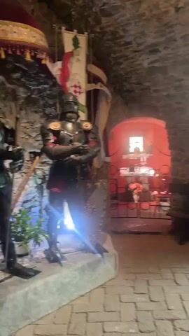The video shows the interior of a stone room, possibly a castle or a historical building. In the foreground, there's a suit of armor standing on a stone platform. Further back, there's a small, red-painted alcove with a religious shrine inside.  The room is furnished with wooden benches and chairs, suggesting it might be a seating area. A table with a potted plant and some floral arrangements is also visible. There are windows with stained glass elements, and the walls are made of rough, uneven stones. The lighting is dim, with light coming in through the windows and some artificial light highlighting the armor.  A flag with a white horse emblem is hung in the background. The floor is paved with bricks.