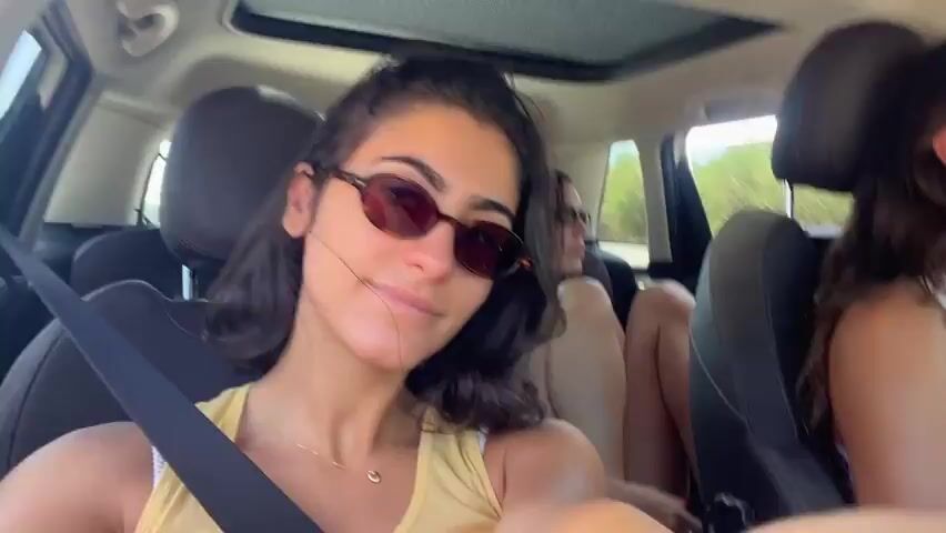 A group of young women are in a car. One woman in the foreground, wearing a yellow shirt and sunglasses, is holding the camera and looking at it. To her right, another woman in a white tank top and sunglasses is driving and gesturing. In the back, a third woman is visible. The car appears to be moving, as indicated by the blurred background and the motion of the passengers. The atmosphere seems casual and upbeat.