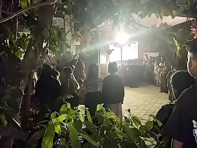 The video shows a crowd of people gathered at night outdoors. There is a bright light illuminating the scene, casting shadows. Some people are standing, while others appear to be sitting. There are trees and foliage in the foreground and background, suggesting an outdoor setting with greenery. The people are dressed in what appear to be traditional clothing. The overall atmosphere seems to be of a gathering or event. The image is blurry and dark due to the nighttime conditions and the quality of the recording.