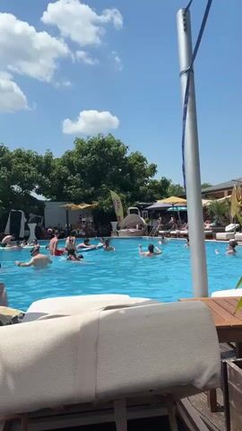 The video shows a busy outdoor swimming pool on a sunny day. Many people are in the water, swimming, floating, and socializing. Some are lying on loungers near the pool, and others are gathered around umbrellas. The pool area is surrounded by trees and buildings, and the sky is blue with some white clouds. There is a tall pole with a blue cable running from it. In the foreground, a portion of a white lounge chair is visible, with a wooden deck and table.