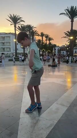 A boy in a light green t-shirt and khaki shorts dances outdoors in a plaza at dusk. He wears blue and gray sandals. Palm trees and buildings line the background. The sky is a mix of orange and pink. Several people walk in the background.