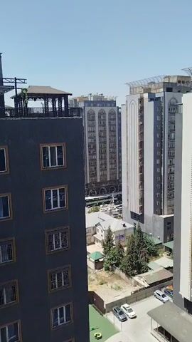 The video shows a high-angle view of an urban environment with multiple buildings and a clear sky. In the foreground, a dark gray building with numerous windows is visible on the left. To the right, there are several taller buildings with a mix of architectural styles and colors, including gray, white, and beige. One of these buildings has a white, curved facade at the top. In the middle ground, there are smaller buildings, some with red roofs, and several trees. A dirt lot with a fence surrounds some of the buildings. A few cars are parked in a lot at the bottom right. The overall impression is a sunny day in a city with a mix of modern and older construction.