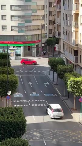 A street scene is shown from an elevated viewpoint. The street is lined with trees and buildings on both sides. There are two cars visible on the road. A red car is seen driving towards the left of the frame, and a white car is moving away from the viewer towards the right. A pedestrian is walking across the street in front of the white car. The road markings indicate parking spots and lanes for traffic. A traffic light is visible in the background, showing a red light. The overall environment appears to be an urban setting during the daytime.