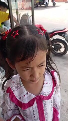 A young girl with pigtails adorned with red star-shaped clips and red bows is shown outdoors. She is wearing a white shirt with a pink floral pattern and pink ruffled trim. In the foreground, she holds a clear plastic bottle of water. The background is somewhat blurred, showing a red motorcycle and what appears to be a food stall with a glass enclosure. The girl looks down at the bottle, then at the camera, and proceeds to drink from the bottle.