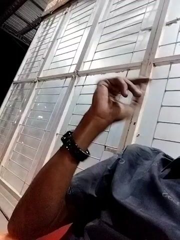 The video shows a person sitting inside a building with their left arm raised towards a window, the hand gesturing as if waving or expressing emphasis.  The window is made of horizontal, white-painted slats with wire mesh covering the gaps.  The person is wearing a dark-colored shirt and a black wristband or watch.  The background shows a building's exterior and a dark sky.  The perspective suggests the camera is being held by the person.