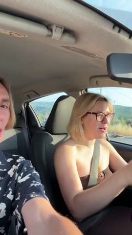 Two people are in a car. The person on the left is a male with blond hair, wearing a dark shirt with a white floral pattern. He is leaning towards the camera and his mouth is open. The person on the right is a female with blond hair and glasses, wearing a strapless brown top. She is looking forward and appears to be driving. Both individuals are wearing seatbelts. The car's interior is visible, including the dashboard and the roof lining. Outside the car, a rural landscape with hills and trees can be seen, with the sun setting and casting a warm light.