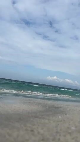 This is a low-angle, eye-level shot of a beach with gentle waves rolling in. The scene is set at Manggis, Indonesia. The water is a striking turquoise color, and the waves create a foamy white surf as they reach the sandy shore. The sand in the foreground is light gray and textured. In the distance, the sea meets the horizon under a sky filled with a mix of white, fluffy clouds and patches of blue. A few boats are visible on the water in the far distance, suggesting a relatively calm day. The lighting indicates it's daytime, likely late morning or early afternoon, with diffused sunlight due to the cloud cover. There are no people visible in the frame, giving the scene a peaceful and serene atmosphere. The overall impression is one of natural beauty and tranquility.