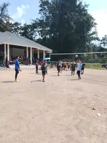 This video shows a group of people playing volleyball on a dirt court. There is a shelter with a roof in the background, where some spectators are sitting. The volleyball net is set up, and several people are actively participating in the game. One person in the foreground is wearing a blue shirt and black shorts, and another person nearby is wearing a black and white shirt and black shorts. The sky is visible with some clouds, and there are trees in the background. The overall scene appears to be an outdoor recreational activity.
