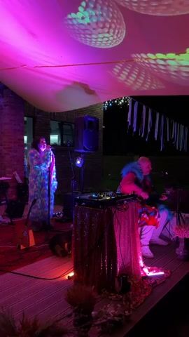 A group of people are on a raised wooden platform outdoors at night. The platform is decorated with pink and gold sequins, as well as some dried plants. There is a fabric canopy overhead with a light projection of what appear to be circular patterns in green and pink. A woman on the left is singing into a microphone while wearing a colorful, furry robe. In the center, a person is seated at a DJ booth with turntables. To the right, another person is playing an instrument, possibly a keyboard. There is a guitar leaning against a stand to the left of the singer. Several speakers and lights are also visible. White streamers hang from above in the background. The overall atmosphere appears to be festive and musical.