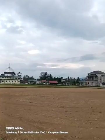 The video shows a rural landscape with a field in the foreground and buildings and mountains in the background. The field appears to be plowed or tilled, with brown soil visible. Several houses and a larger, multi-story building are situated in the middle ground, surrounded by trees. In the distance, mountains are visible under a cloudy sky. The video pans slowly from left to right, offering a panoramic view of the scene. A watermark at the bottom of the screen indicates the recording was made on June 26, 2026, at 17:40, in Kabupaten Breuen, using an OPPO A6 Pro phone. No people or animals are clearly visible in the frame, and no specific actions or interactions are depicted. The environment suggests a daytime scene in a rural area, possibly agricultural land.