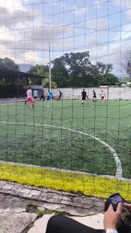 The video shows a group of people playing soccer on a green artificial turf field, seen through a black net. The players are wearing different colored shirts and shorts. There is a white wall behind the field, with some trees visible beyond the wall. The sky is cloudy.