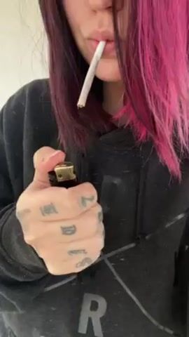 A person with dyed pink hair is shown from the shoulders up wearing a dark gray hooded sweatshirt. They are lighting a cigarette in their mouth using a black and gold lighter held in their tattooed left hand. After lighting the cigarette, they take a puff and exhale a large cloud of smoke.