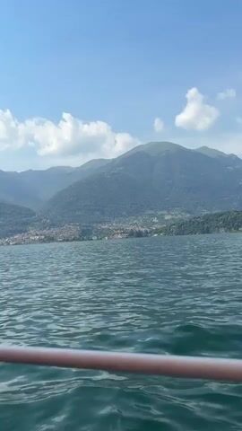 This video captures a scenic view of a body of water, likely a lake, with a distant town nestled at the base of lush green mountains. The water is a deep, shimmering green, with gentle waves rippling across its surface, suggesting a calm and serene atmosphere. In the foreground, a reddish-brown railing runs horizontally across the bottom of the frame, indicating the viewpoint is from a boat or a similar elevated structure on the water.

The sky above is a vibrant blue, dotted with fluffy white clouds that drift lazily, hinting at pleasant weather, possibly midday or early afternoon. The mountains, covered in dense foliage, rise majestically in the background, their slopes creating a dramatic backdrop for the town. The town itself appears to be a collection of buildings with red-tiled roofs, typical of many European villages, and it is situated along the shoreline, giving the impression of a picturesque lakeside community.

The overall mood is one of tranquility and natural beauty. The scene evokes a sense of peaceful travel or leisure, perhaps a boat trip on a sunny day. There is no visible text in the media. The setting strongly suggests a location like Chiesa, Italy, known for its beautiful lakes and mountainous terrain.