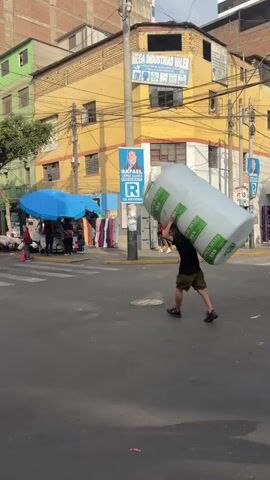 The video shows a busy street scene in what appears to be a city in Latin America, evidenced by the architecture and signage. A man is carrying a large roll of what seems to be mattress padding on his shoulder, walking across the street. Another man follows him, carrying a black plastic bag. The street is active with pedestrians, street vendors under blue umbrellas, and parked motorcycles. Buildings of varying heights and colors flank the street. Political posters featuring a man's image can be seen on the side of the buildings. Overhead, there is a network of power lines. The sky is overcast, and the lighting is muted.