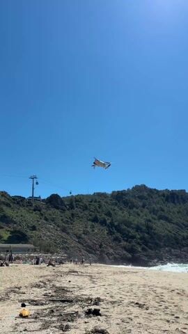 The video shows a white drone flying in a clear blue sky above a beach. The beach has a sandy area littered with debris, and people are seen sunbathing and walking around. In the background, there is a green, hilly area, and a cable car system is visible to the left. The drone moves closer to the camera.