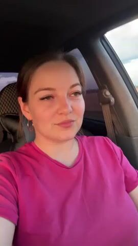 A woman with brown hair and green eyes, wearing a bright pink t-shirt and hoop earrings, is sitting in the driver's seat of a car. She is looking directly at the camera and smiling. The camera then pans to the right, showing the woman's hand on the steering wheel as she drives. The car is moving along a road, with fields of crops and a cloudy sky visible through the windows. The interior of the car is visible, including the dashboard, steering wheel, and rearview mirror.